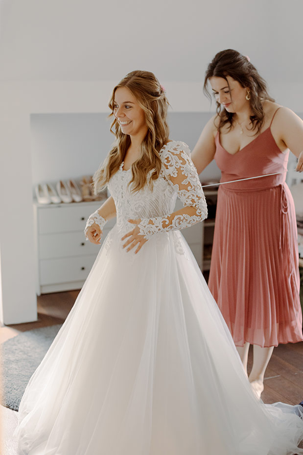 Bride in a white lace wedding gown with long sleeves, smiling as a bridesmaid adjusts her dress in a bright room with a dresser in the background.