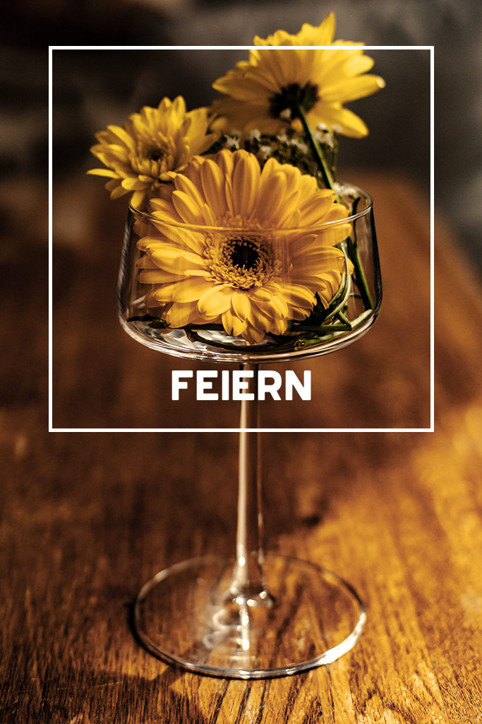 Yellow flowers in a wine glass on a wooden table, overlaid with the word FEIERN, conveying a celebratory theme.