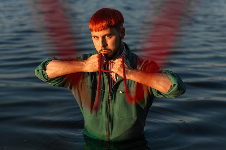 Man in a green coverall stands waist-deep in water, pulling red straps toward his face with a serious expression, red light trails across the scene.