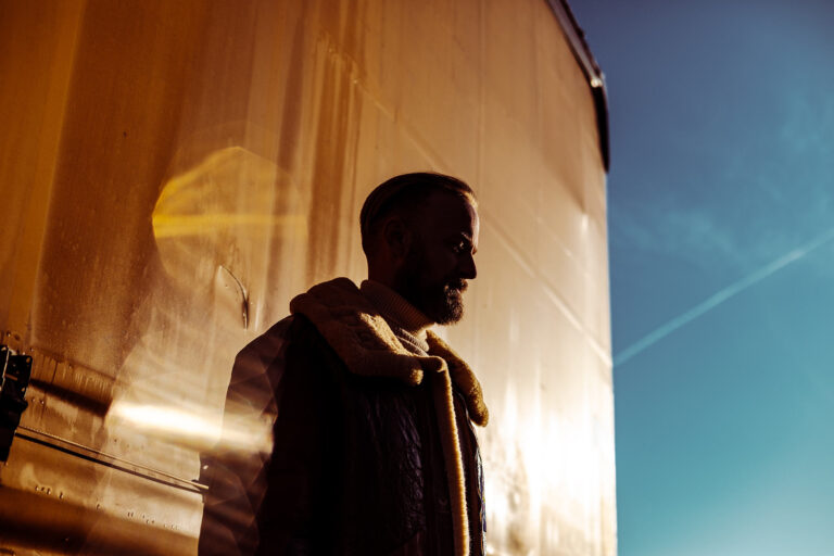 Silhouette of a bearded man in a warm jacket standing beside a sunlit orange train car against a blue sky.