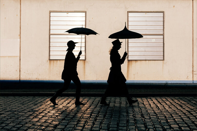 Silhouettes of a man and a woman walking with umbrellas along a cobblestone street, backlit by a pale wall with windows.