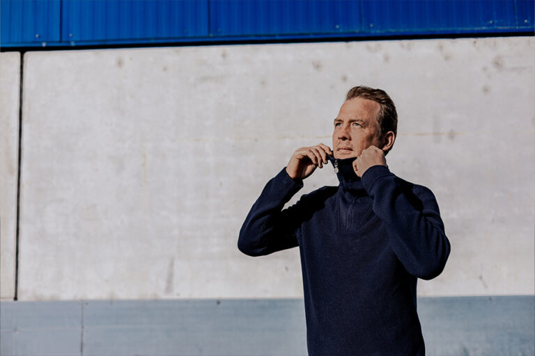 Man in a dark blue sweater zips up the collar outdoors against a light concrete wall with a blue top edge.