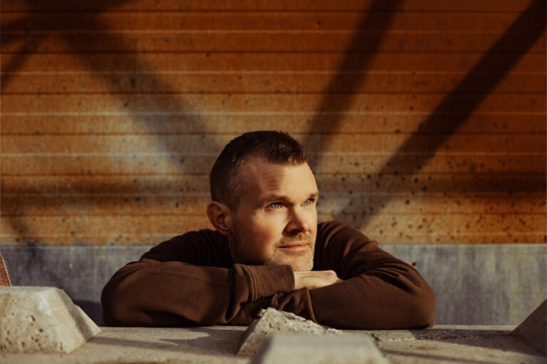 Portrait of a man in a brown sweatshirt resting his arms on a concrete barrier, gazing off to the side against a brick wall backdrop.