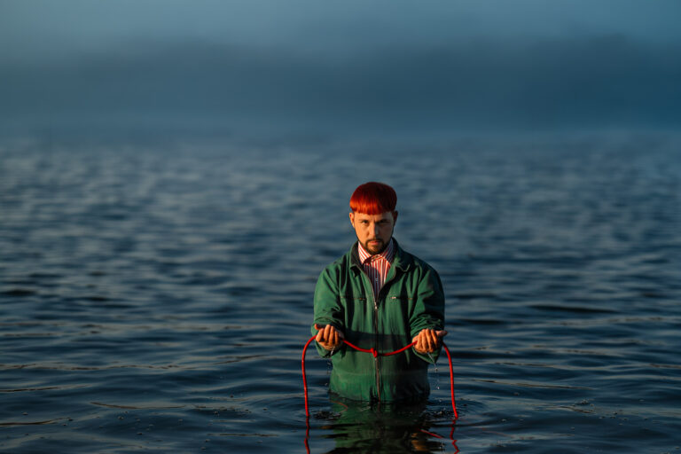 Person standing chest-deep in water, red-haired, wearing a green jacket, holding a red rope that extends into the water.