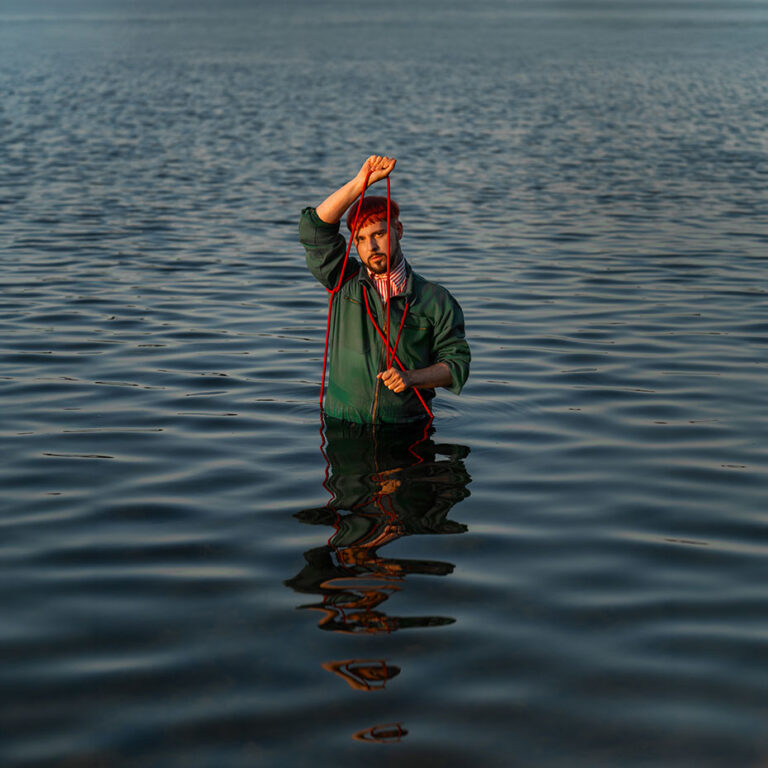 Man waist-deep in calm lake water, lifting a red rope above his head with a green jacket, reflection visible on the rippling surface.